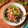 Black bean quinoa bowl topped with avocado, pico de gallo, cilantro, and creamy hummus-lime dressing served in a white bowl.