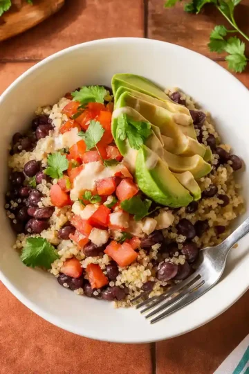 Black bean quinoa bowl topped with avocado, pico de gallo, cilantro, and creamy hummus-lime dressing served in a white bowl.