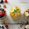 A vibrant flat lay of three anti-inflammatory overnight oats variations in glass jars. Toppings include mixed berries and walnuts, tropical mango with coconut, and banana with chocolate, all beautifully arranged on a rustic white wood table.