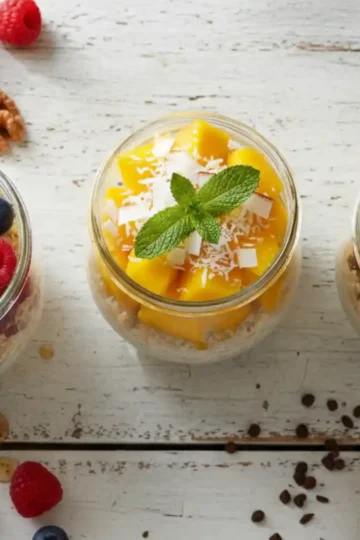 A vibrant flat lay of three anti-inflammatory overnight oats variations in glass jars. Toppings include mixed berries and walnuts, tropical mango with coconut, and banana with chocolate, all beautifully arranged on a rustic white wood table.
