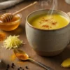 A warm ceramic mug filled with steaming golden milk (Haldi Doodh) on a rustic wooden table, surrounded by turmeric, honey, grated ginger, and black pepper, glowing softly in natural morning light.